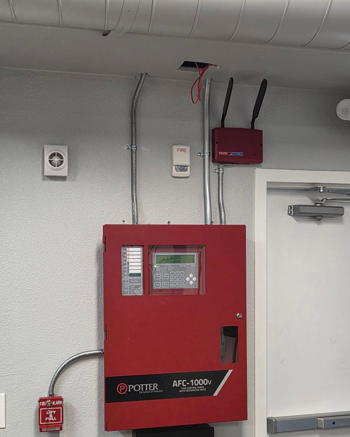 Red fire alarm control panel on a gray wall with conduit, a strobe and a manual pull station