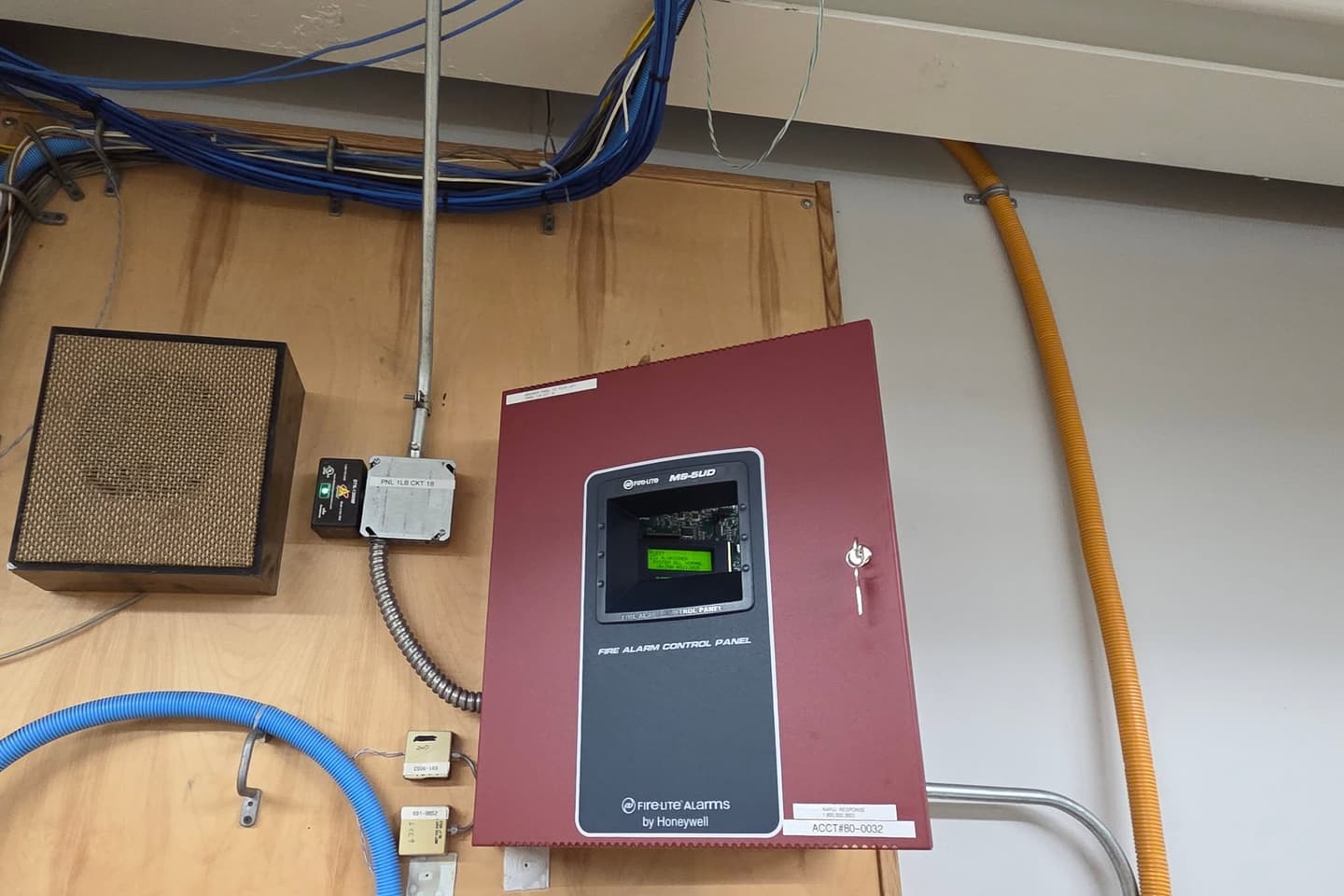 Fire alarm control panel mounted on a plywood backboard beside an older speaker box